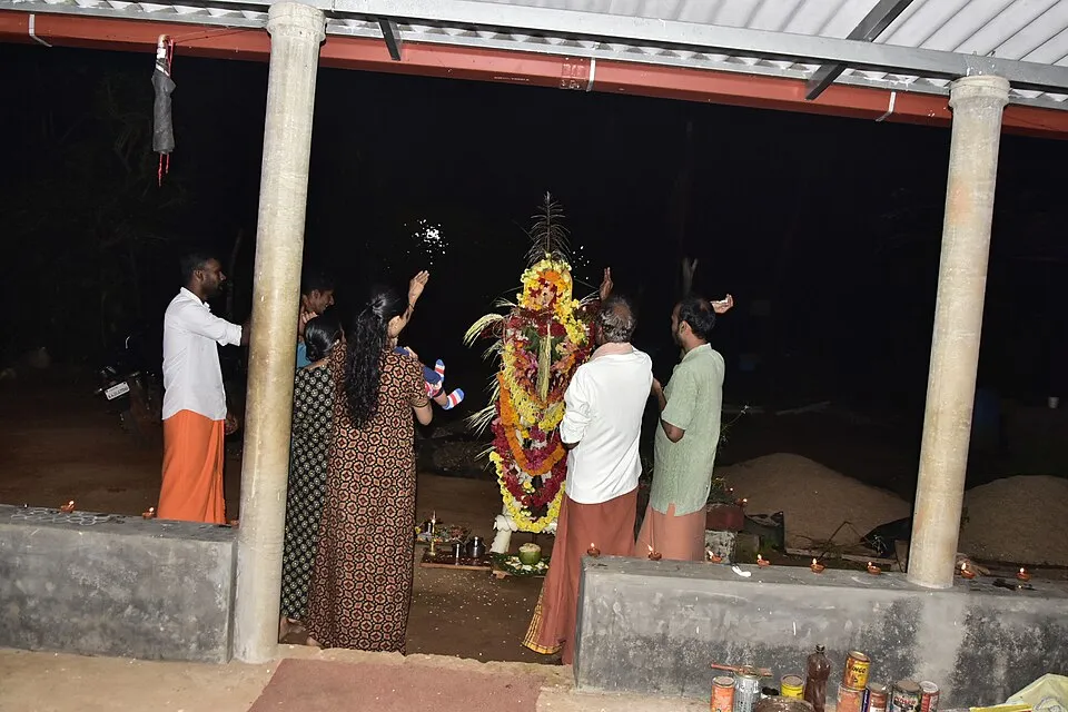 A family praying at night before a tall flower-decked pole set up for Bali, with small lamps around it
