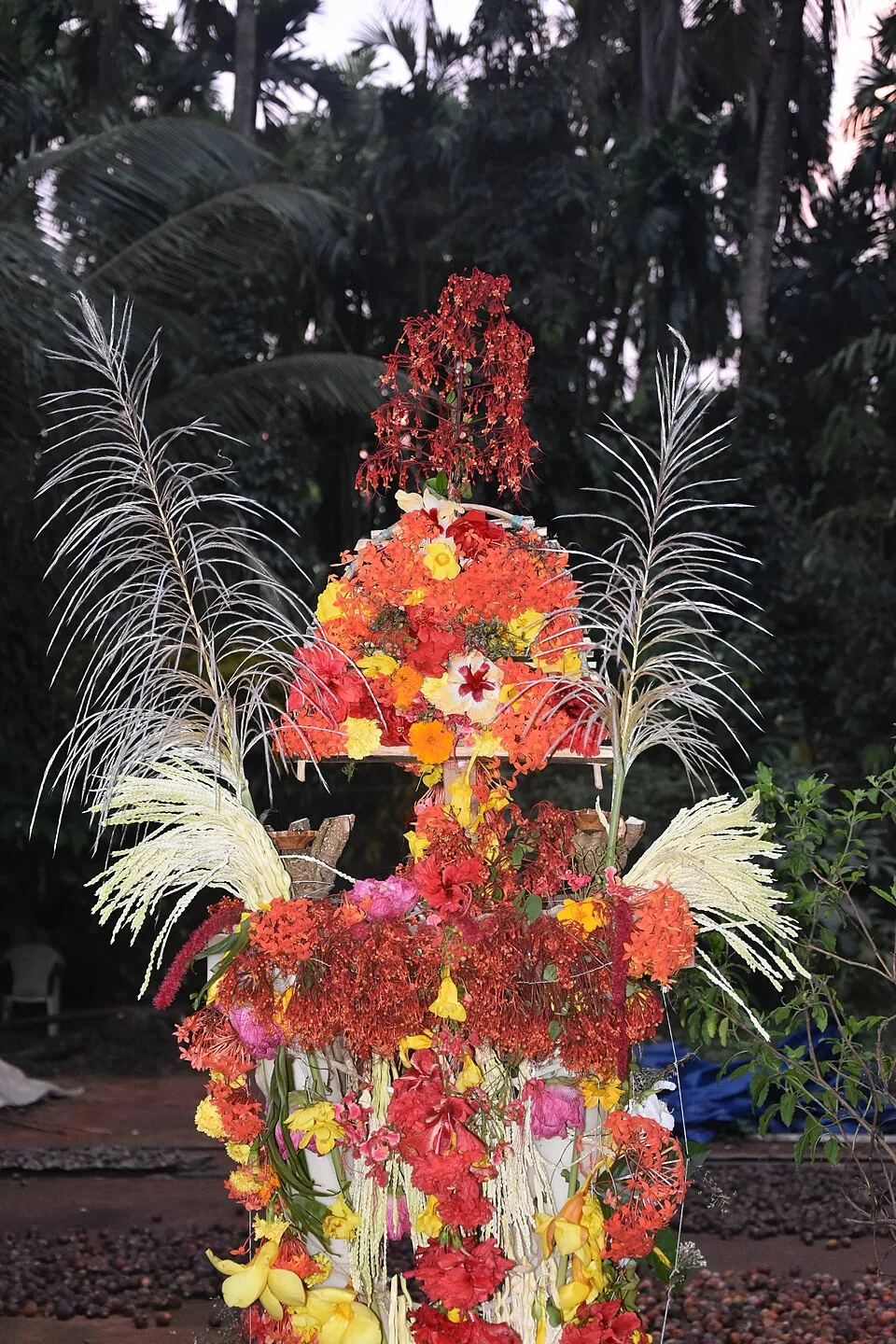 A tall pole thickly decorated with red, orange and yellow flowers and white palm blossoms, set up for Bali Padyami