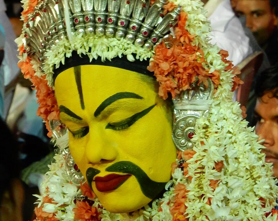 Close-up of a performer as Bobbarya, face painted yellow with a thick black moustache and red lips, under a silver crown and heavy jasmine and orange garlands