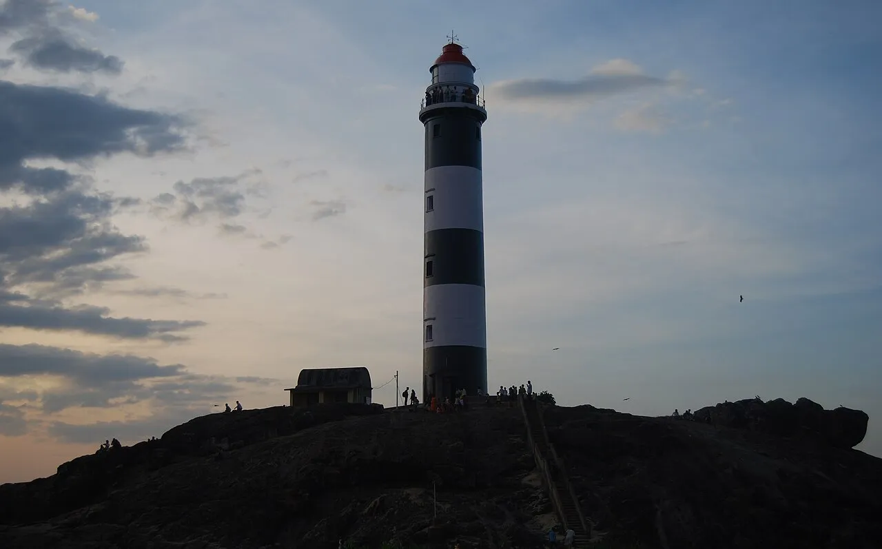 The striped lighthouse at Kaup on a dark rocky hill against an evening sky, with small figures of visitors around its base