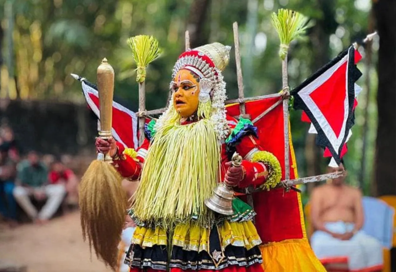 A performer as Bobbarya at a kola, face painted yellow under a silver crown, in a skirt of tender palm leaves, holding a long brass-capped staff and a bell before red and white flags