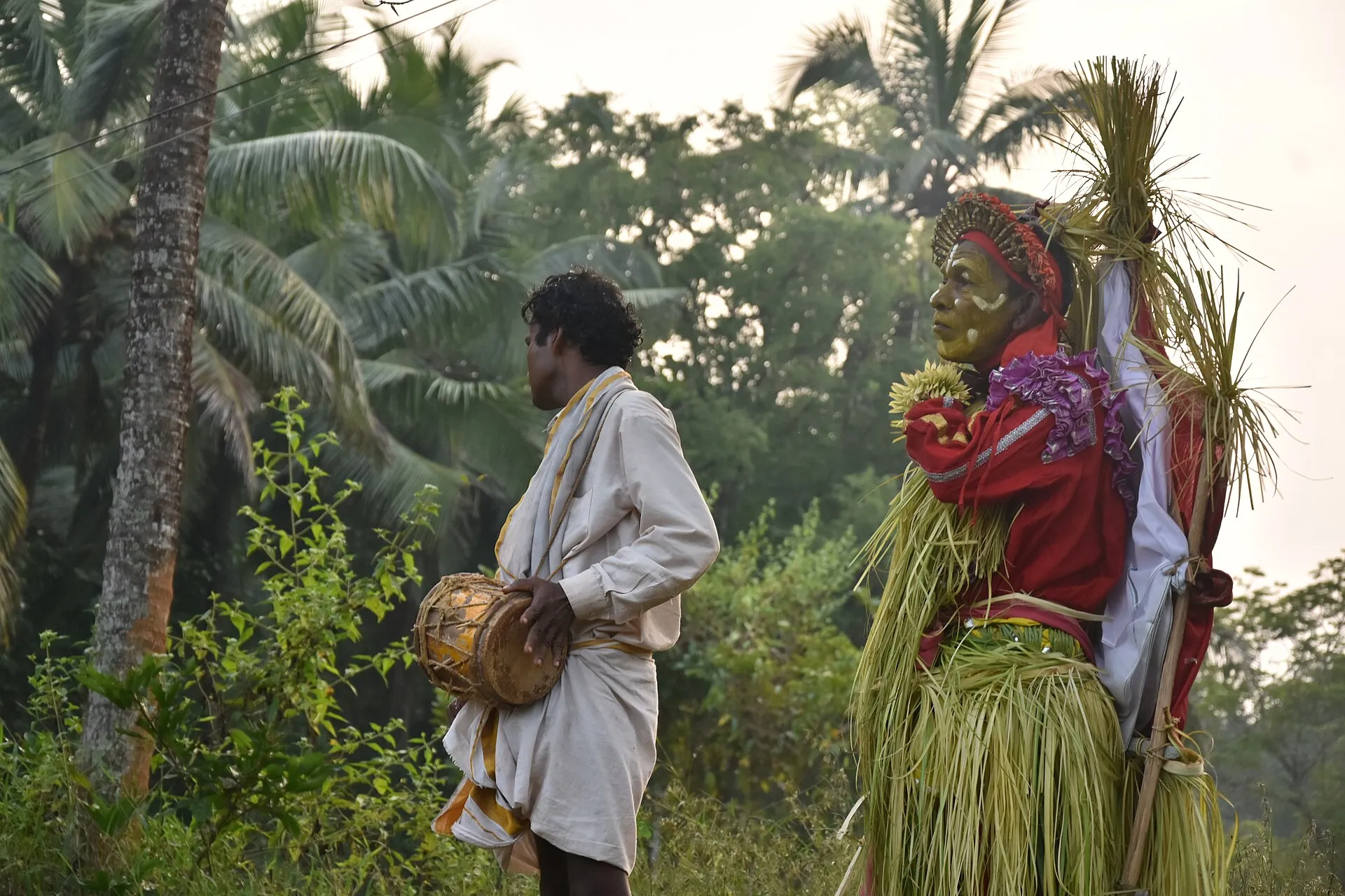 A performer dressed as the spirit Urave, in a palm-leaf skirt, beside a drummer in a field