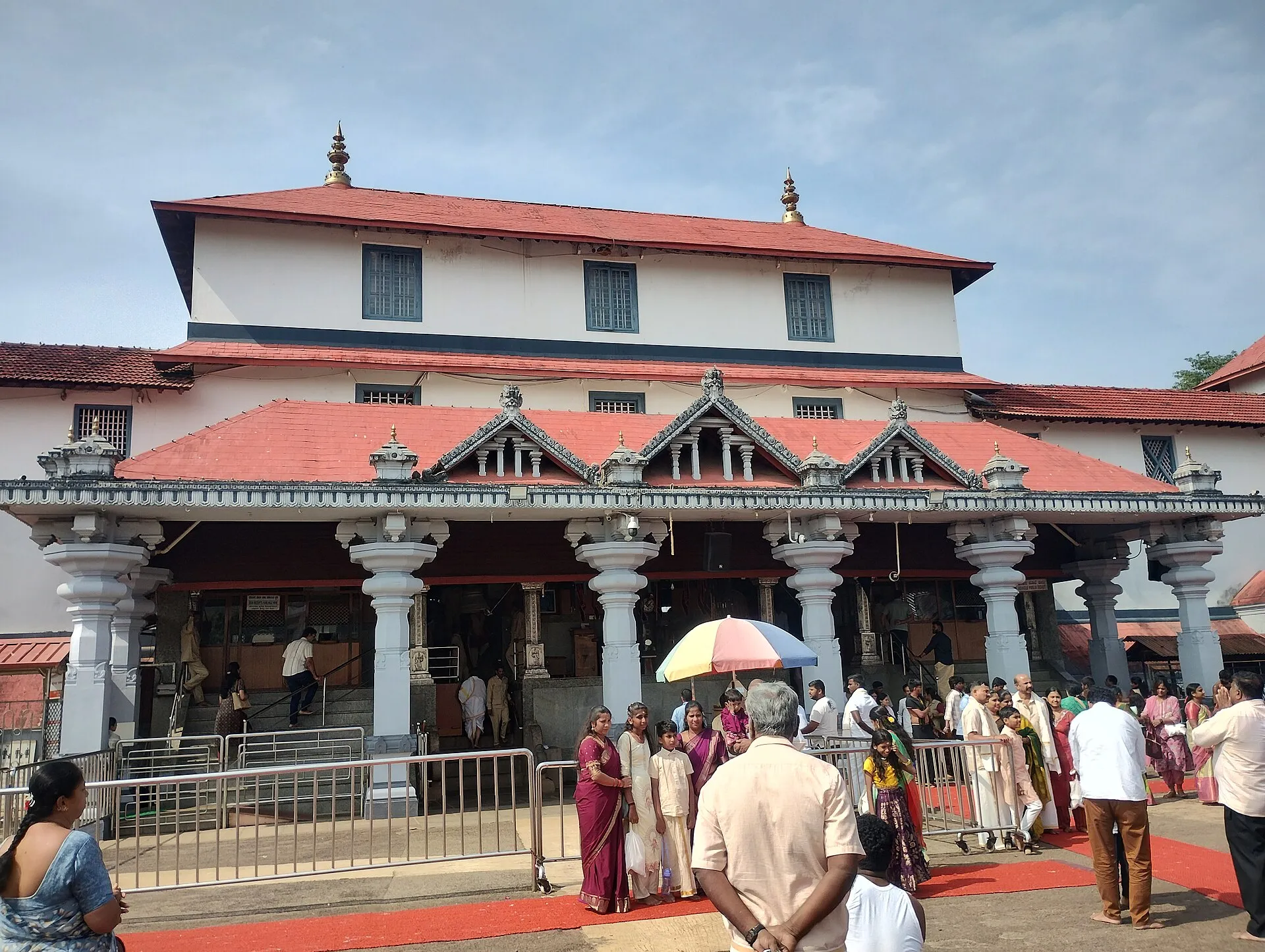 The front of the Manjunatha temple at Dharmasthala, white walls and red-tiled roofs above a pillared porch, with pilgrims queueing in front
