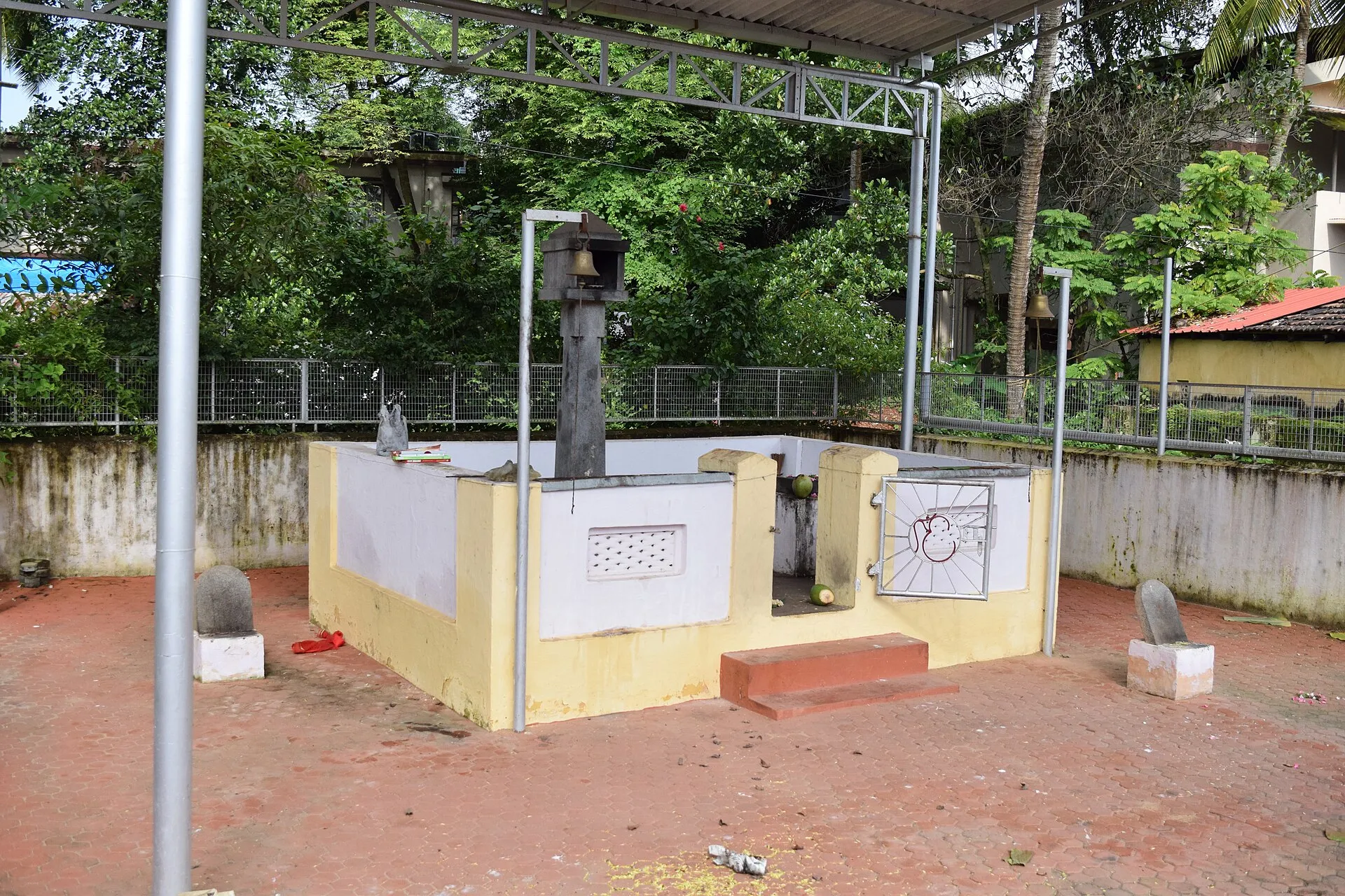 A low walled platform shrine painted yellow and white, with a bell on a post and small upright stones on pedestals around it, under a metal canopy