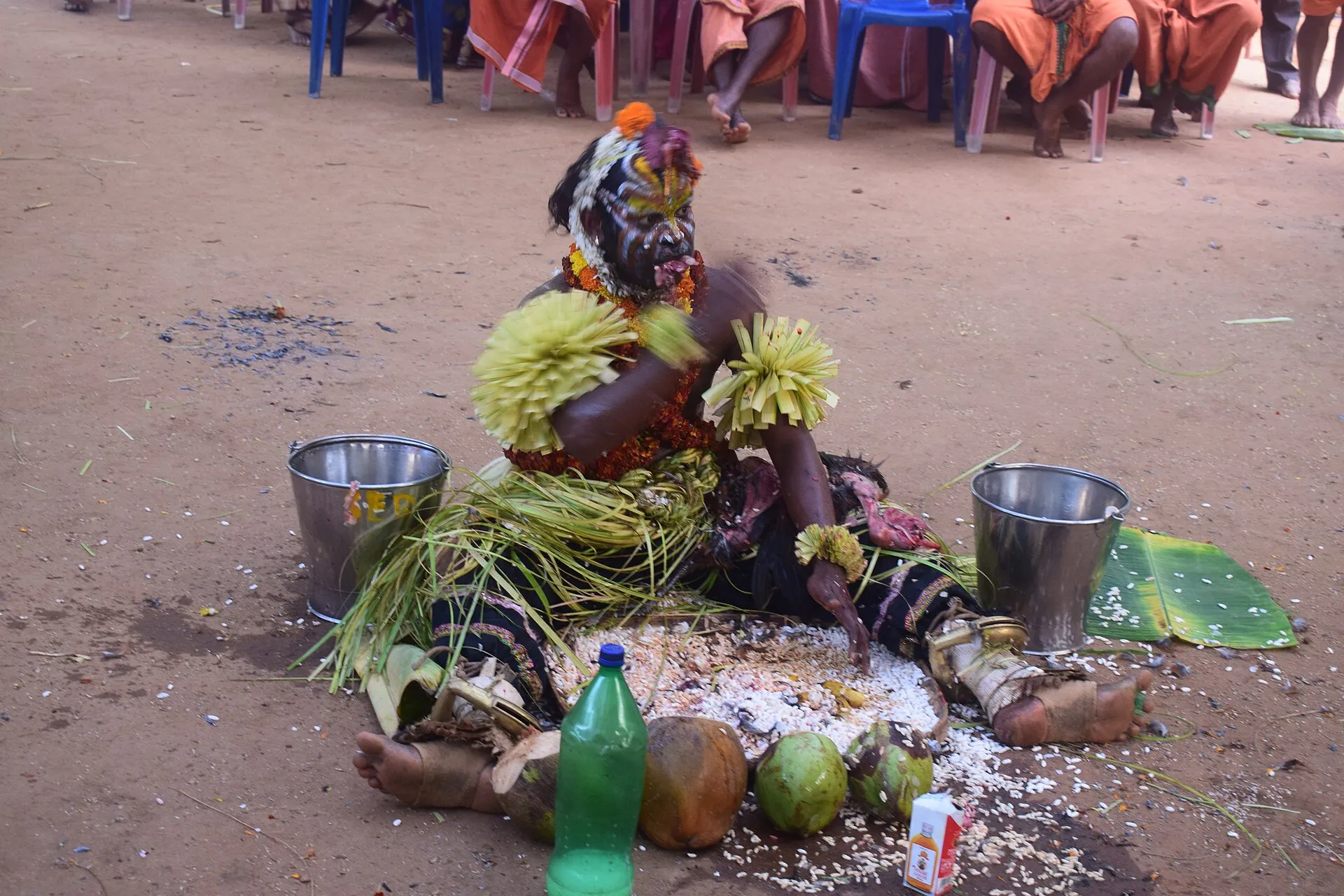 A performer in palm-leaf costume and painted face sits on the ground before heaps of rice and coconuts, with metal buckets on either side and people watching