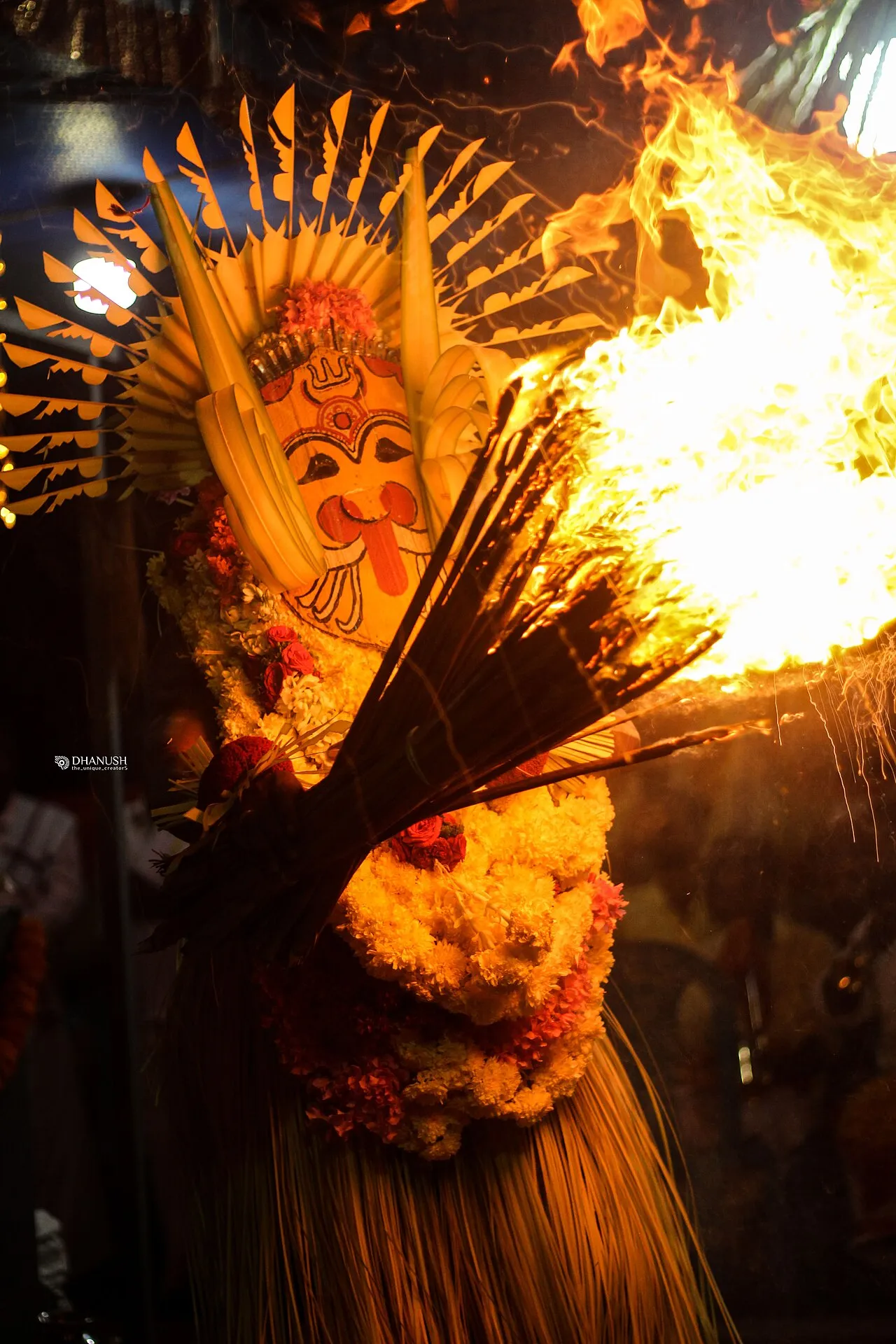 A performer in a tall headdress and a painted mask with a protruding tongue, garlanded with flowers, holding a blazing torch of palm leaves