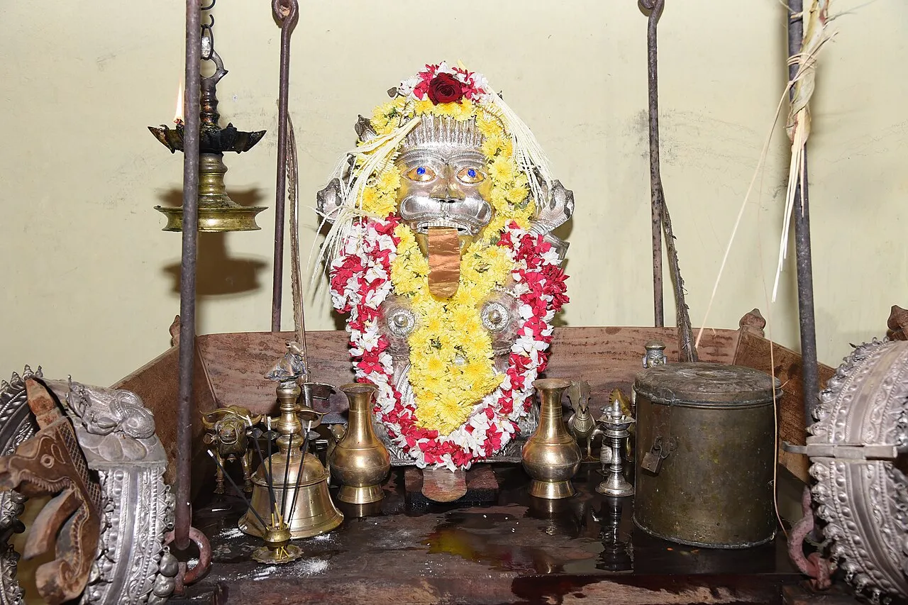 A silver Jumadi mask garlanded with yellow and red flowers in a shrine, with brass lamps, bells and vessels around it