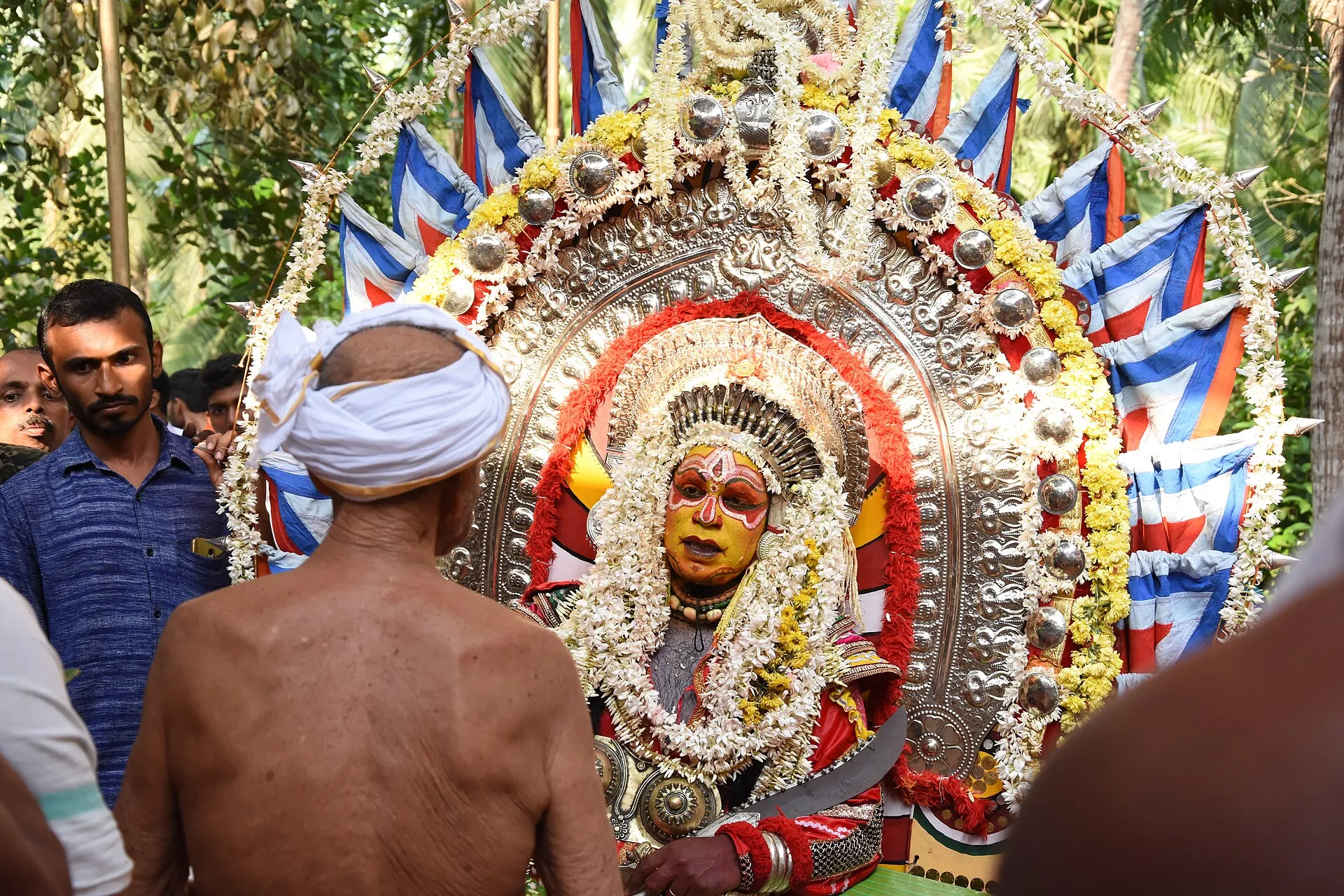 A performer as Jumadi at a kola, face painted yellow and red, before a great silver ani hung with flowers and flags, with men watching