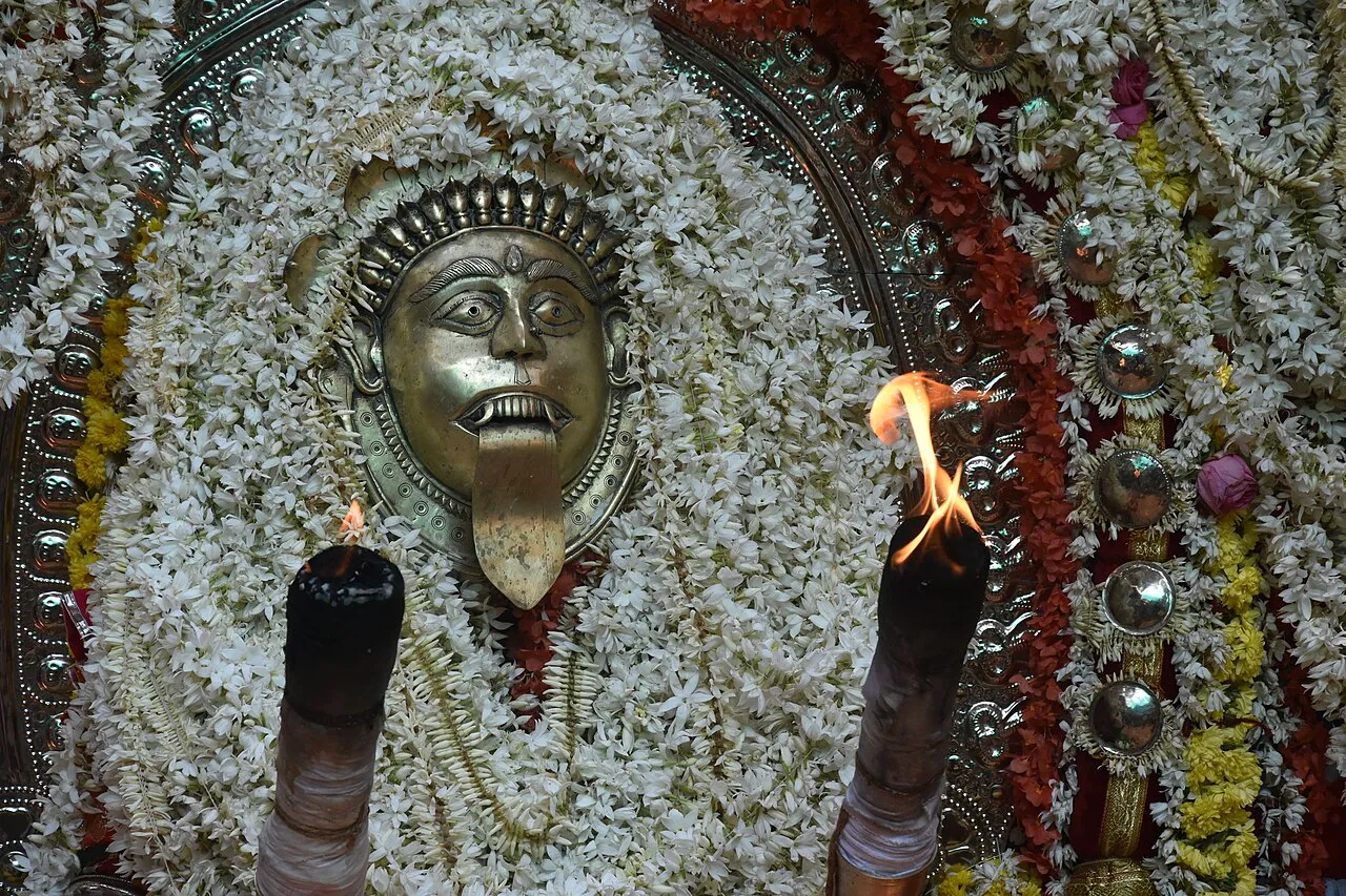 A metal Jumadi mask with a long protruding tongue, set in jasmine garlands between two burning torches