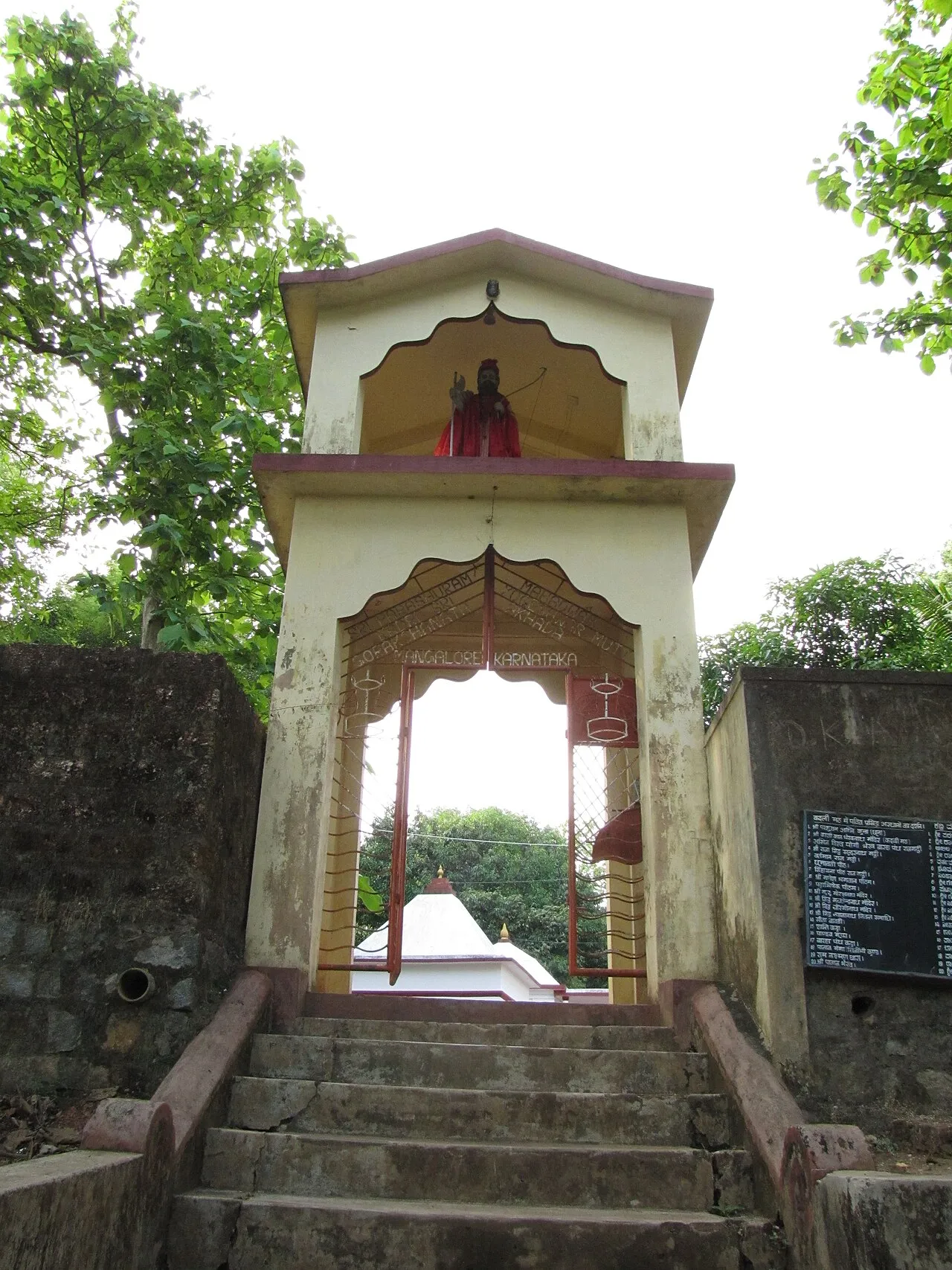 A pale arched gateway with a small shrine on top, at the head of stone steps among trees, leading to the Jogi Math