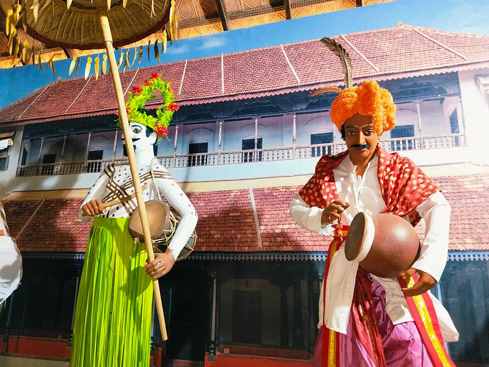 Model figures of Kangilu performers in a display: one in a green fringe costume with a flower headdress, a drum and a palm-leaf umbrella, beside a figure holding a pot drum