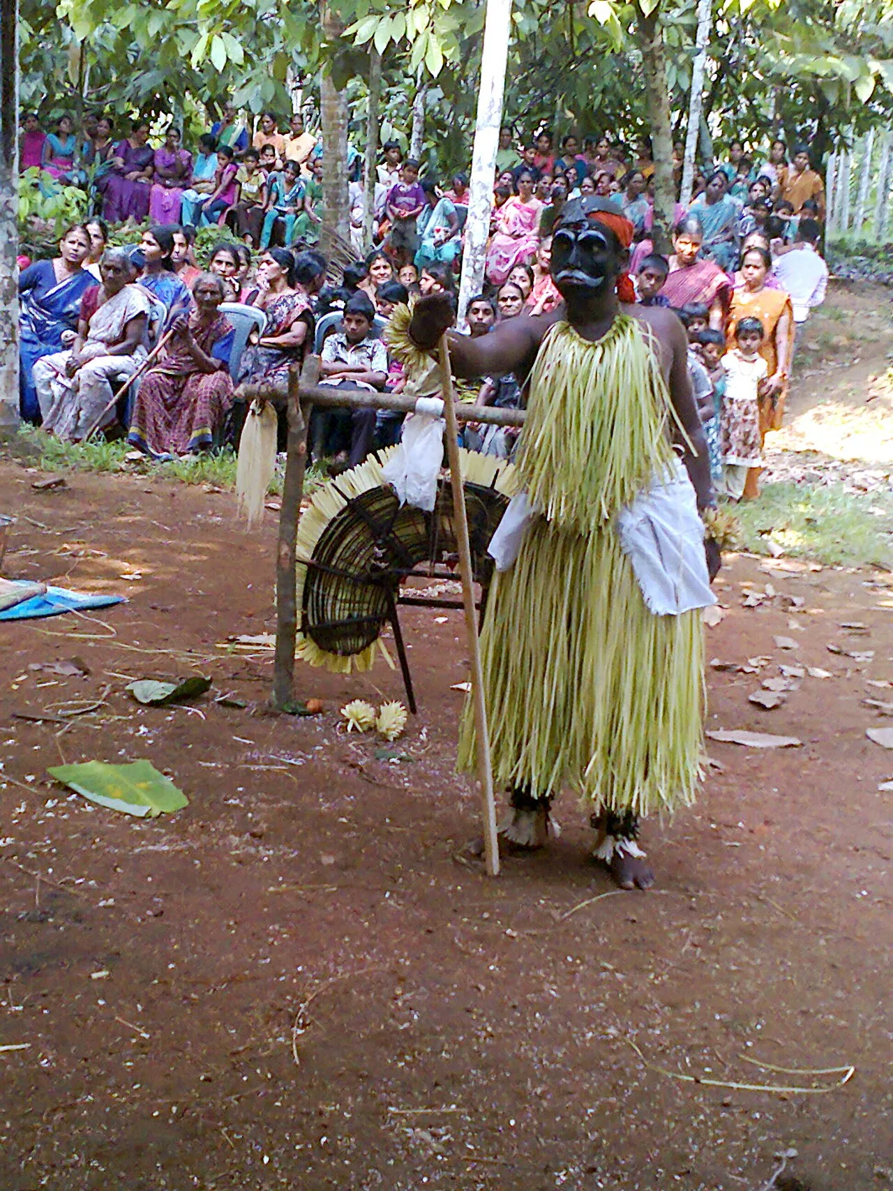 A performer as Koragajja in a skirt of pale palm leaves, holding a staff, with villagers seated under trees behind him