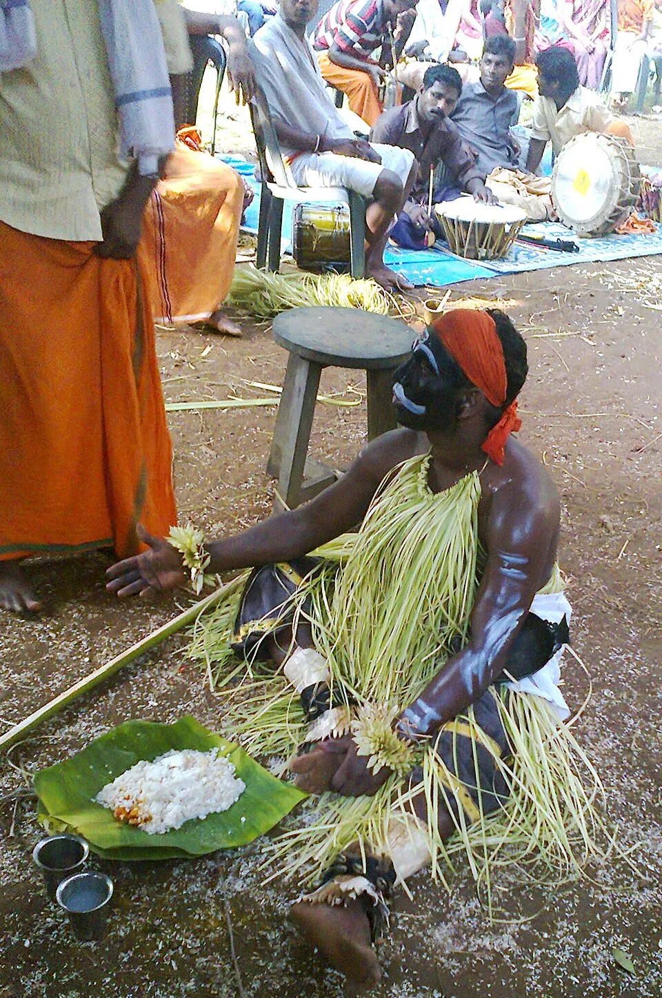 A performer as Koragajja, seated on the ground in a palm-leaf skirt, reaching towards rice served on a plantain leaf