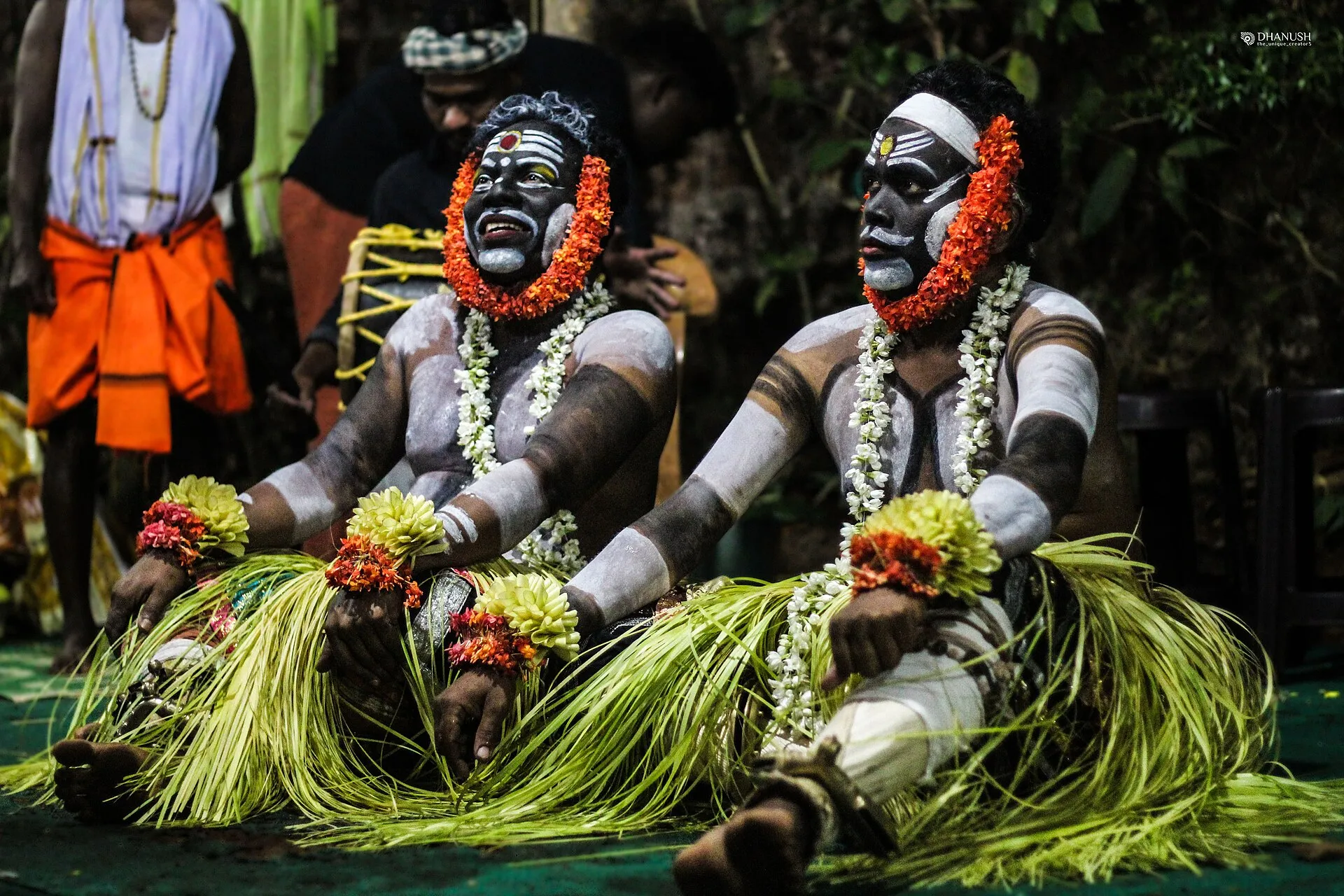 Two performers as Koragajja sit side by side, faces painted black and white, in skirts of tender palm leaves and garlands of orange flowers