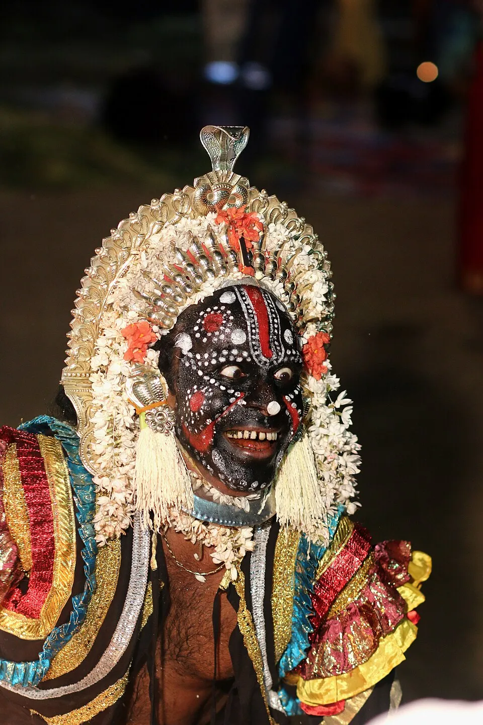 Close-up of a performer as Kalkuda, his face painted black and covered in white dots with red lines, under a silver crown edged with jasmine and red flowers