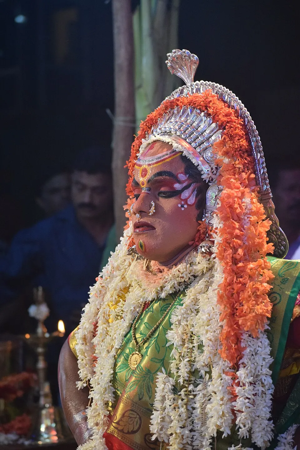 Close-up of a performer as Kallurti, face painted pink with red and white patterns, under a silver crown and thick garlands of orange and white flowers, with a brass lamp burning behind