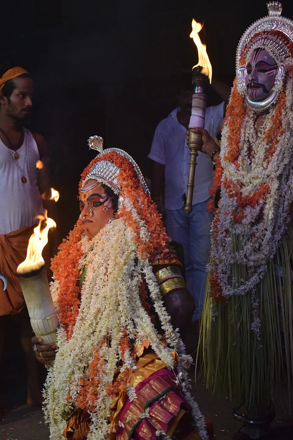Kallurti, bent forward under a silver crown and heavy jasmine and orange garlands, holds a burning torch at a night kola, while Kalkuda, his face painted grey-blue, stands tall behind her with another torch