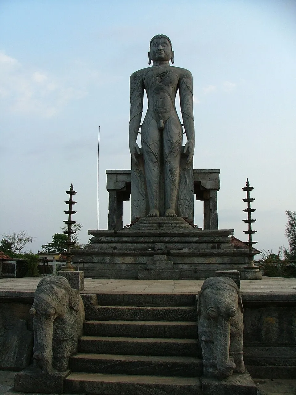 The tall stone statue of Bahubali at Venur, standing on a high stepped platform guarded by two stone elephants, between two slender lamp pillars