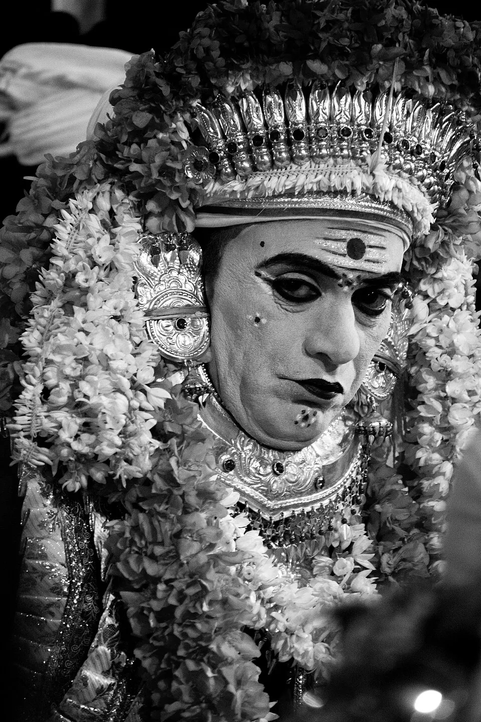 Black and white close-up of a performer as Kodamanithaya, with lined eyes and a dot on the forehead, under a tall ornate crown and heavy garlands