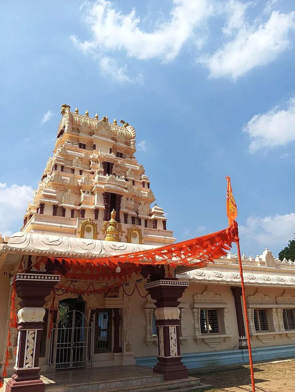 The front of the Kodamanithaya shrine at Thodar, a cream and maroon building with a tall carved tower, strung with orange bunting beside a flagpole