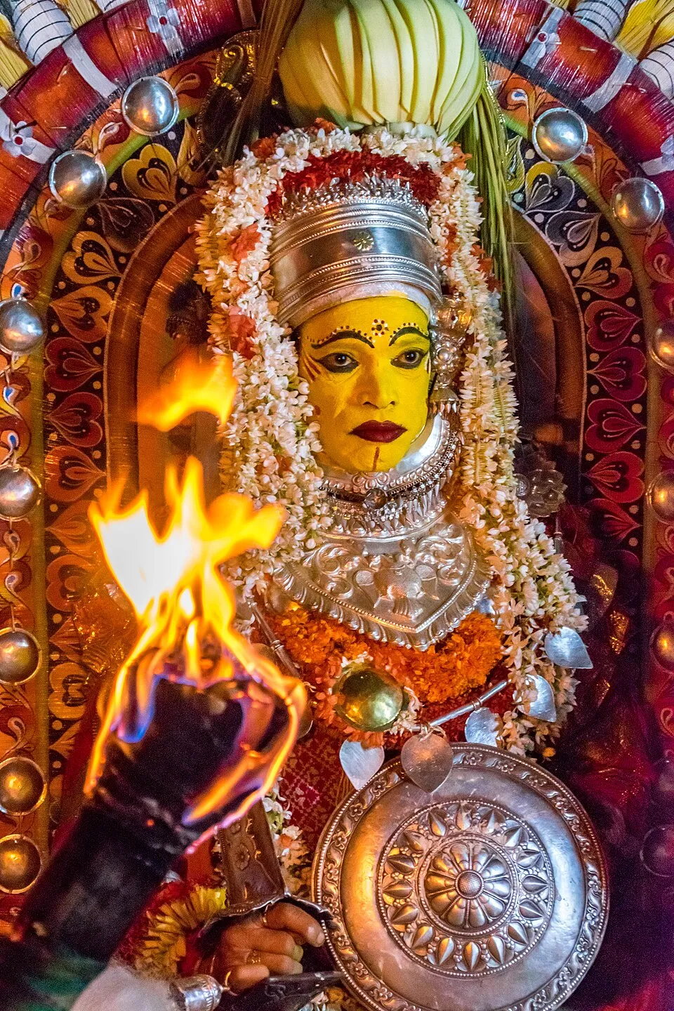 Kodamanithaya at a nema, face painted yellow under a silver headdress and jasmine garlands, holding a silver shield, with a blazing torch held up before him
