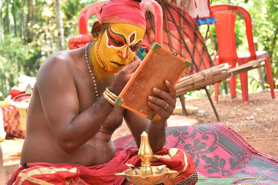 A performer painting his face yellow and red before a kola, using a hand mirror