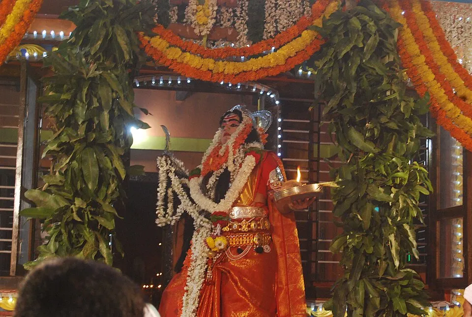 A performer in garlands and an orange silk costume, holding a lamp, at a Koti-Chennaya nema in a decorated shrine