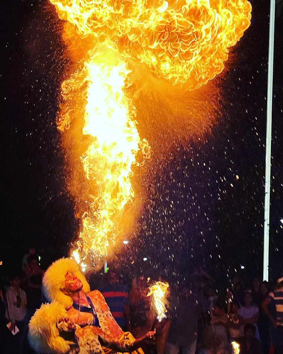 A tiger dancer breathing a column of fire at night, with a crowd watching