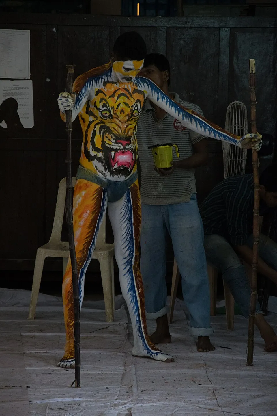 A dancer painted from head to foot in tiger stripes, standing with a stick in each hand while the paint dries