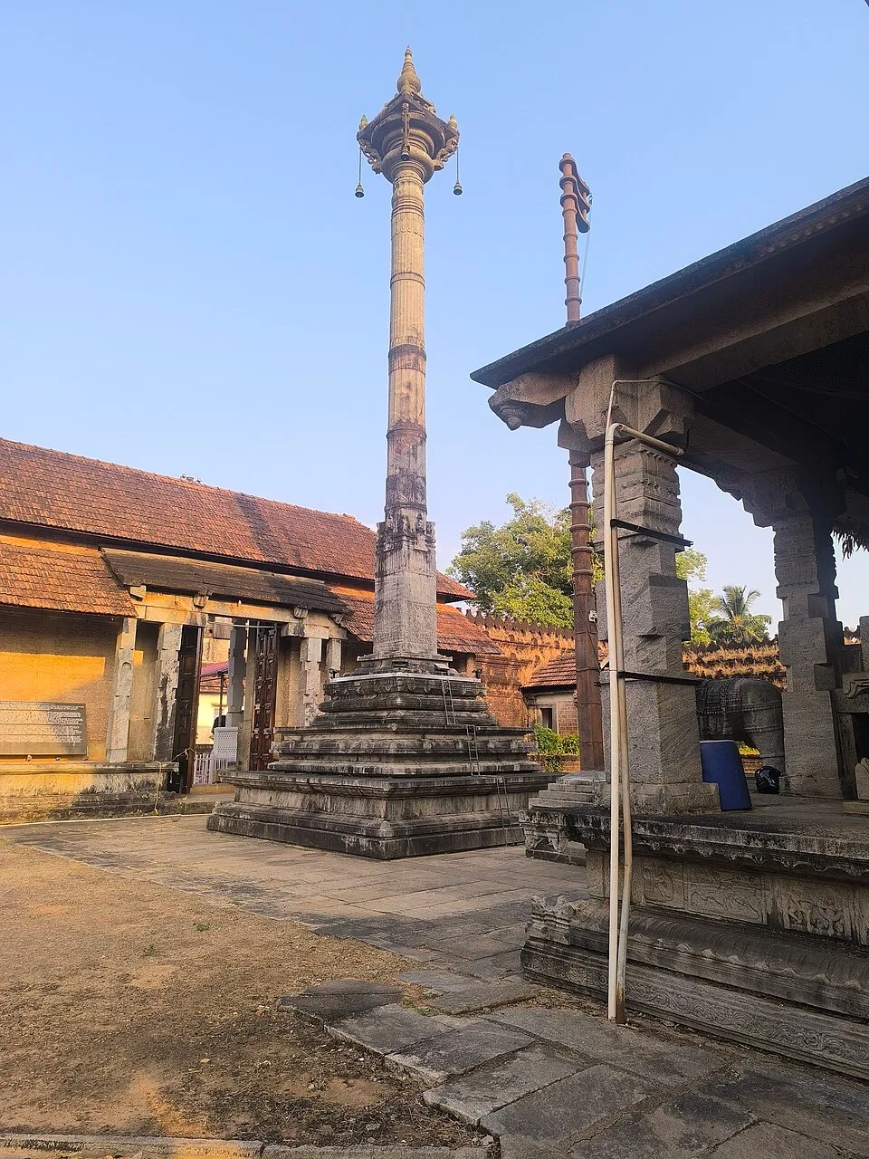 The tall slender stone manastambha in front of the Thousand Pillar Basadi, rising from a stepped base against a blue sky
