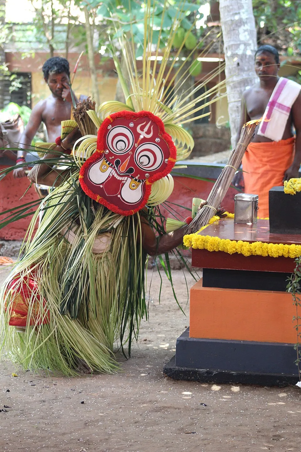 A performer as Maisandaya, bent low in a costume of tender palm leaves, wearing a large painted red and white mask, beside a garlanded shrine platform