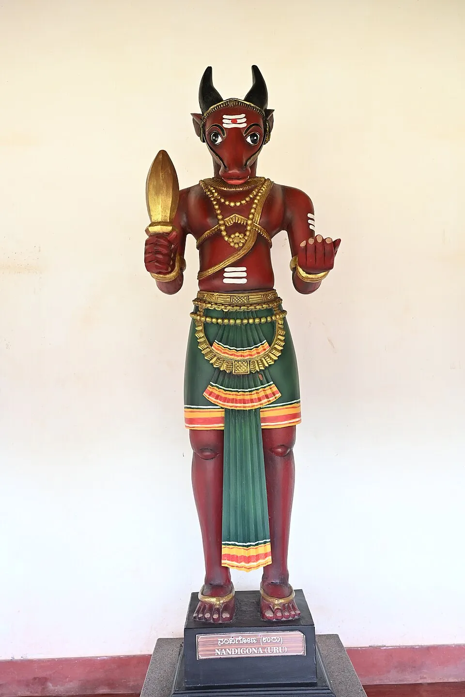 A painted wooden figure of Nandigona with a bull's head and horns and a man's body, holding a short golden sword, on a black pedestal