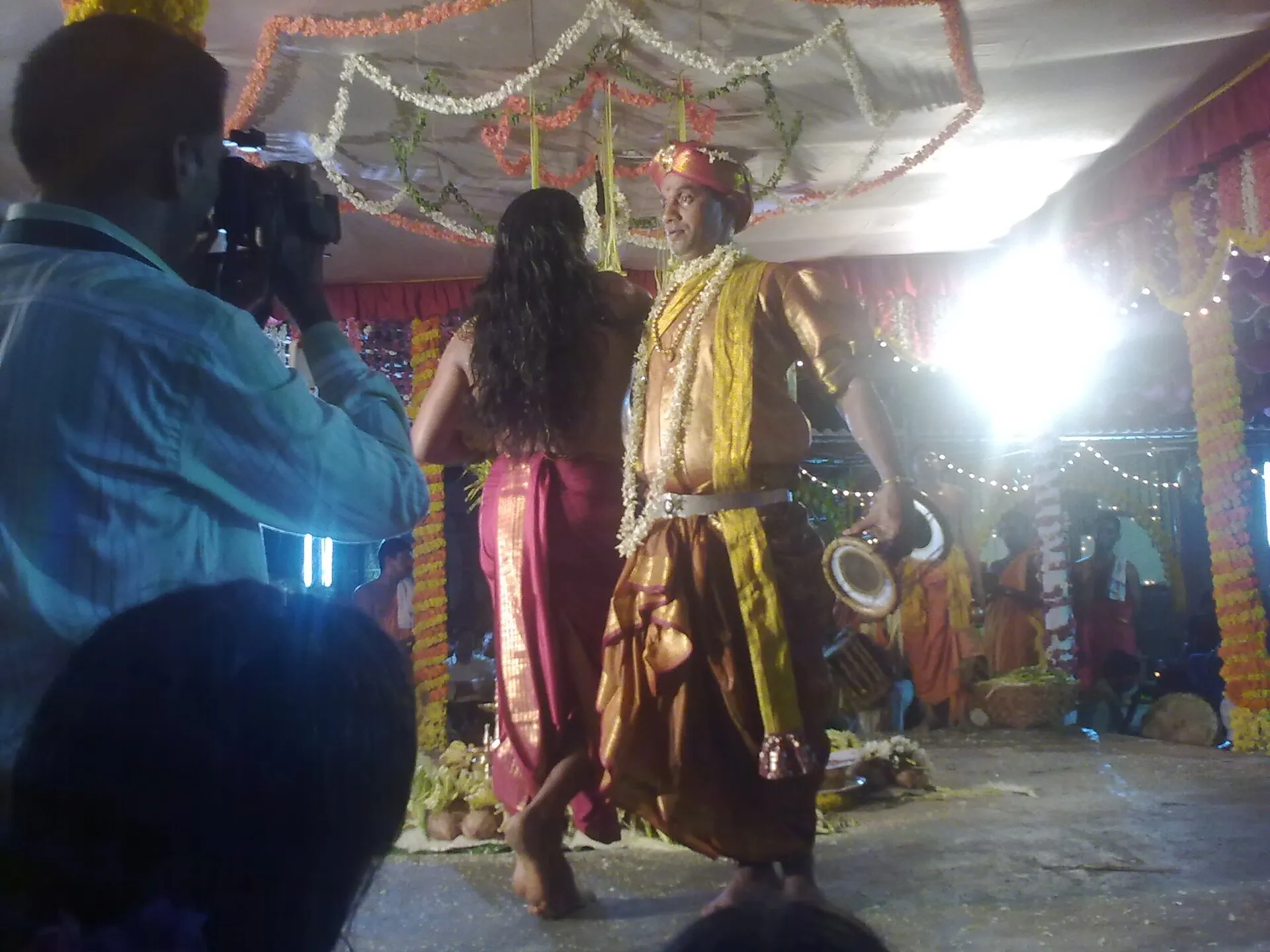 Under a canopy of flower garlands, a performer in gold holds a small hourglass drum while a second performer with long loose hair, dressed in a red sari, moves beside him