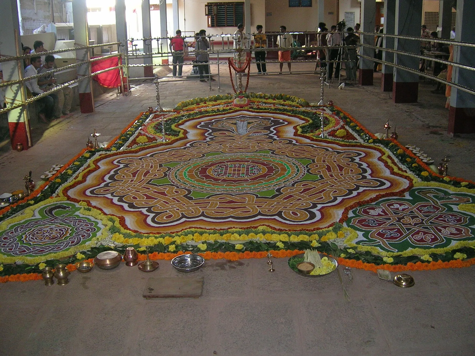 A large serpent mandala drawn on the floor in coloured powders, with knotted coils, framed by flowers and brass ritual vessels