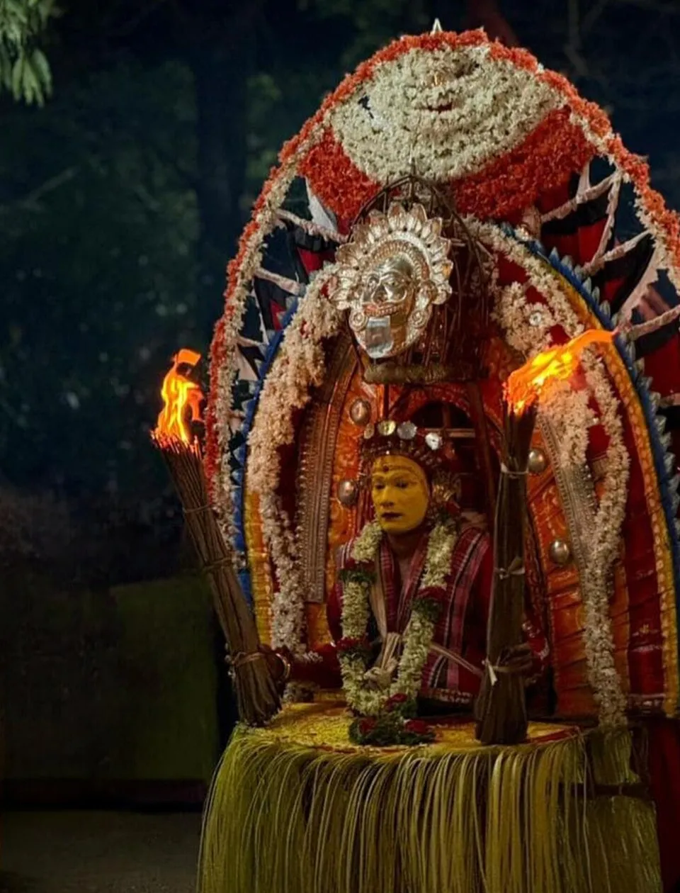 Pilichamundi seated before a great arched ani of flowers and cloth with a silver mask at its top, between two blazing torches of dried palm leaf