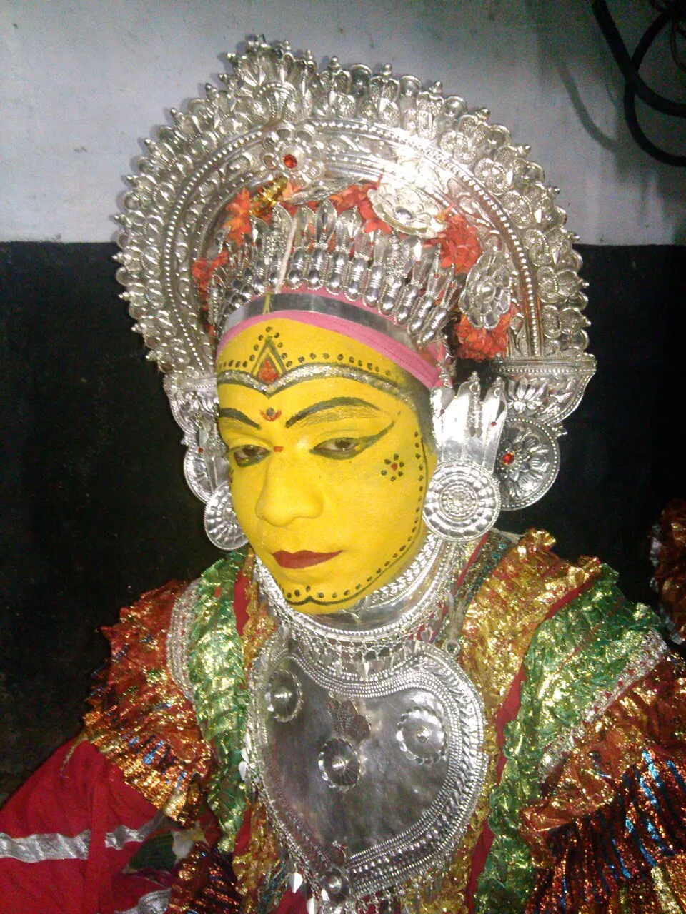 A performer as Pilichamundi, face painted yellow with fine black dotted lines, wearing a tall silver crown and a silver breastplate