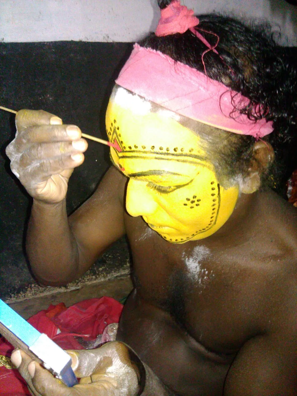 A performer painting fine lines onto his yellow face with a thin stick, looking into a hand mirror, before becoming Pilichamundi