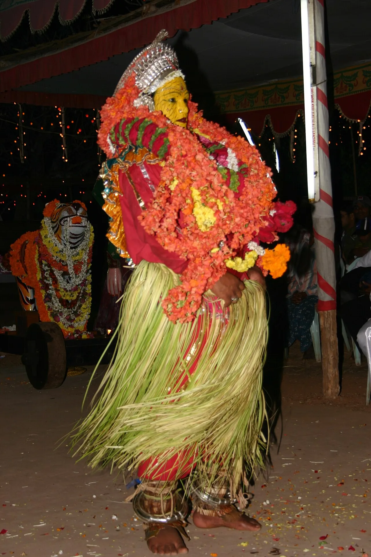 A performer as Pilichamundi at a night kola, face painted yellow, heavy with orange flower garlands and a skirt of tender palm leaves, with a garlanded wooden tiger on a cart behind