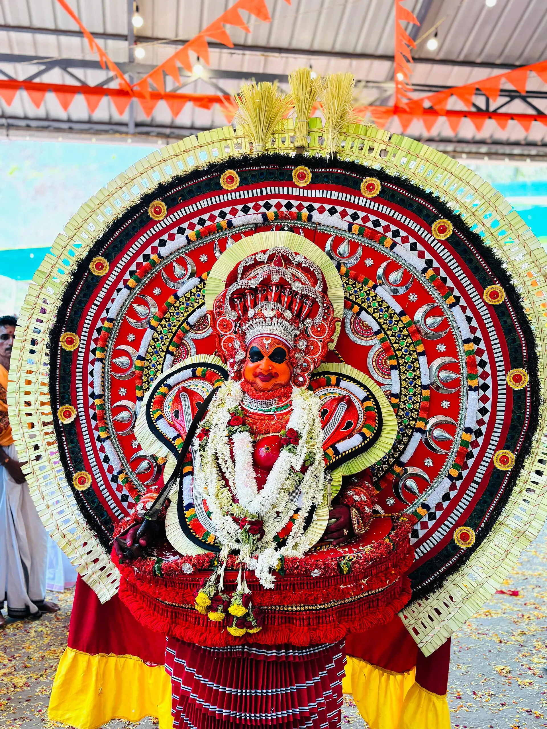 A performer as Rakteshwari at a kola, in red and gold with a great round ani of flowers and cloth