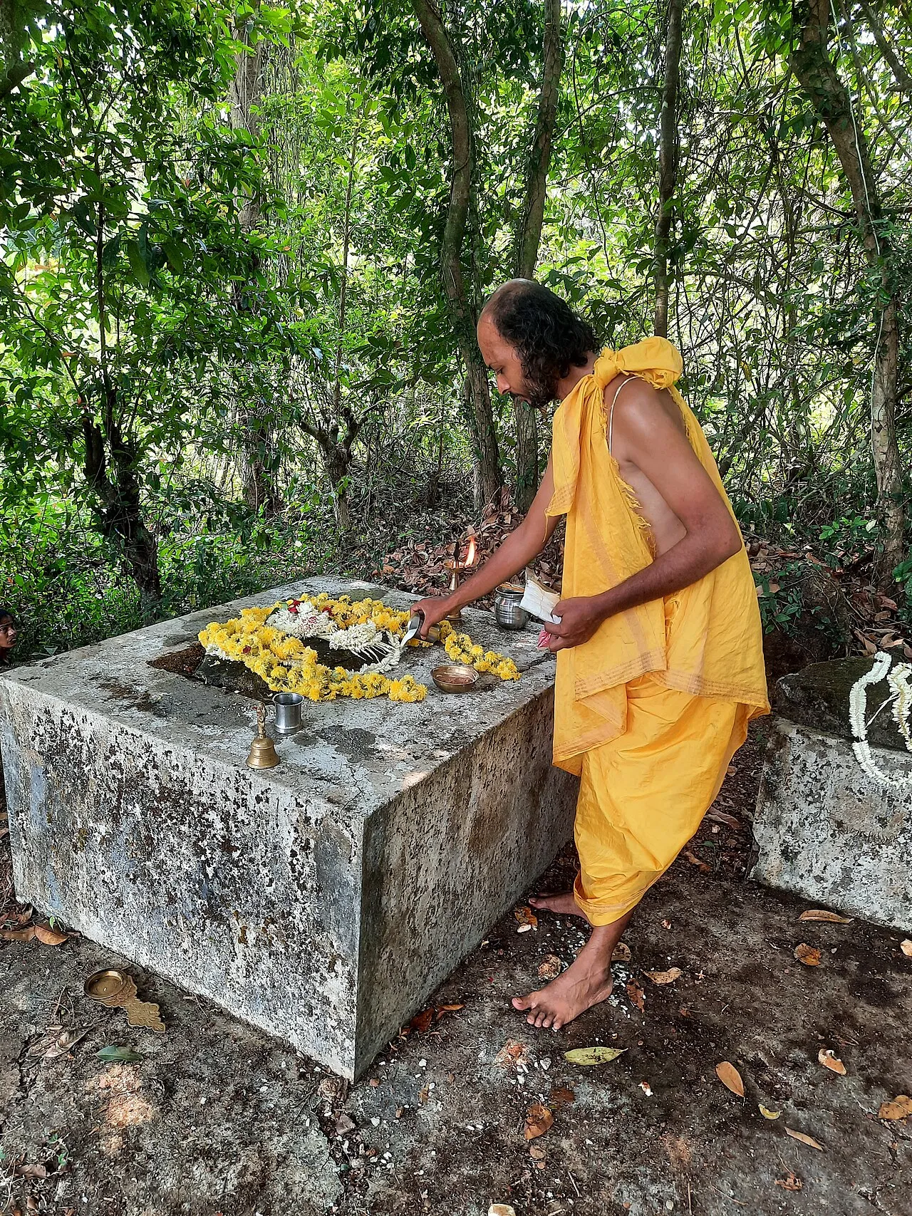 A man lays offerings on a stone platform among forest trees at a Lekkesiri ceremony