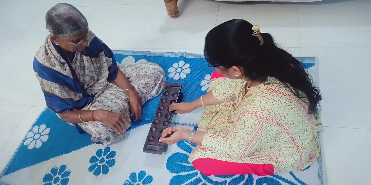An older woman and a younger woman sit on a mat playing chenne mane, a long wooden board with two rows of hollows