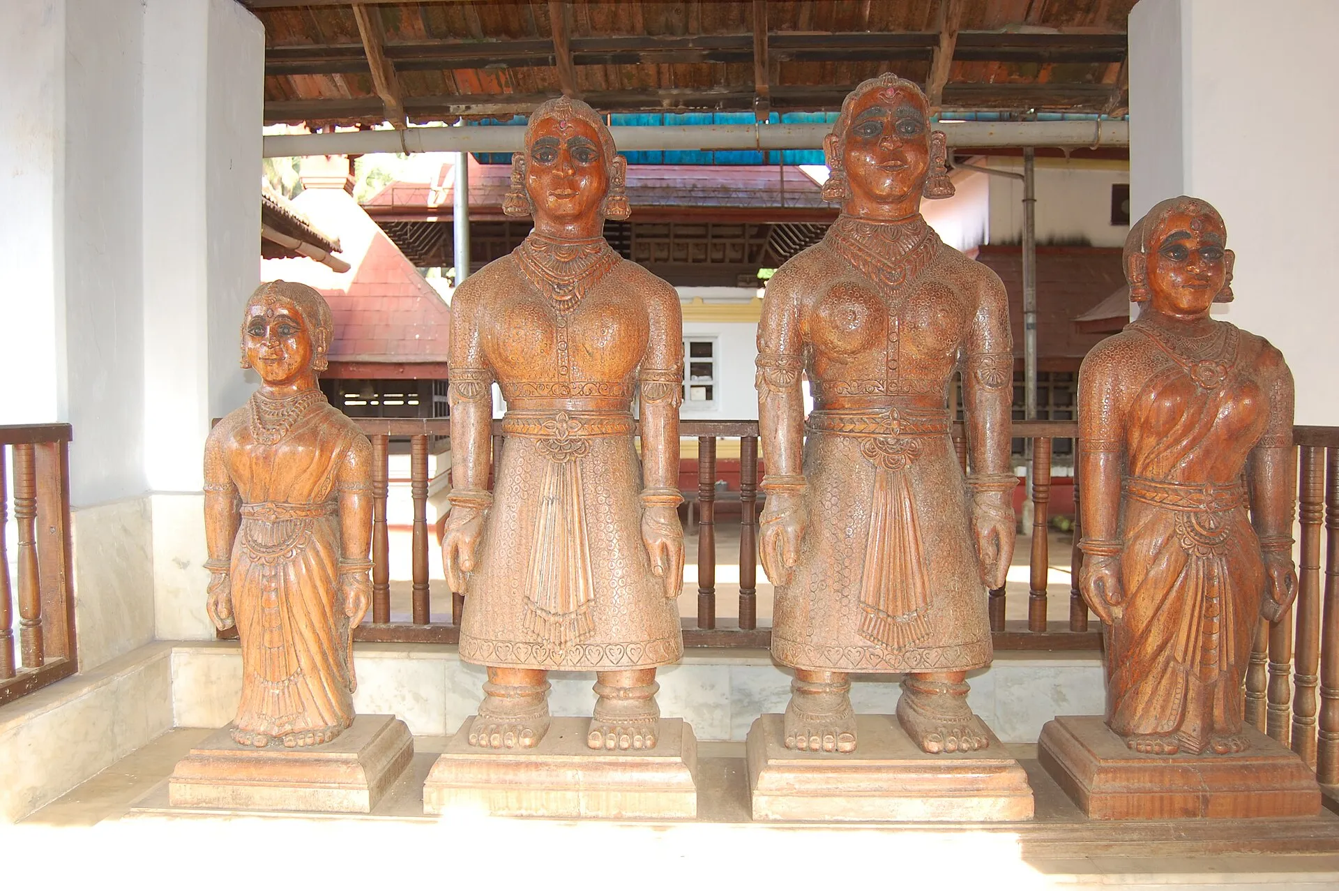 Four carved wooden figures of women stand side by side on low pedestals in a temple hall