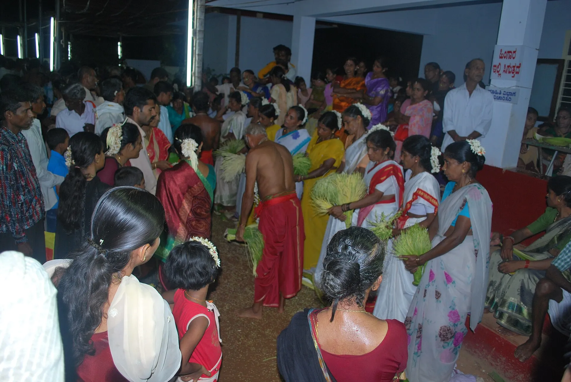 Women in white saris, holding green sheaves, stand in a crowd at night around a bare-chested priest in a red cloth