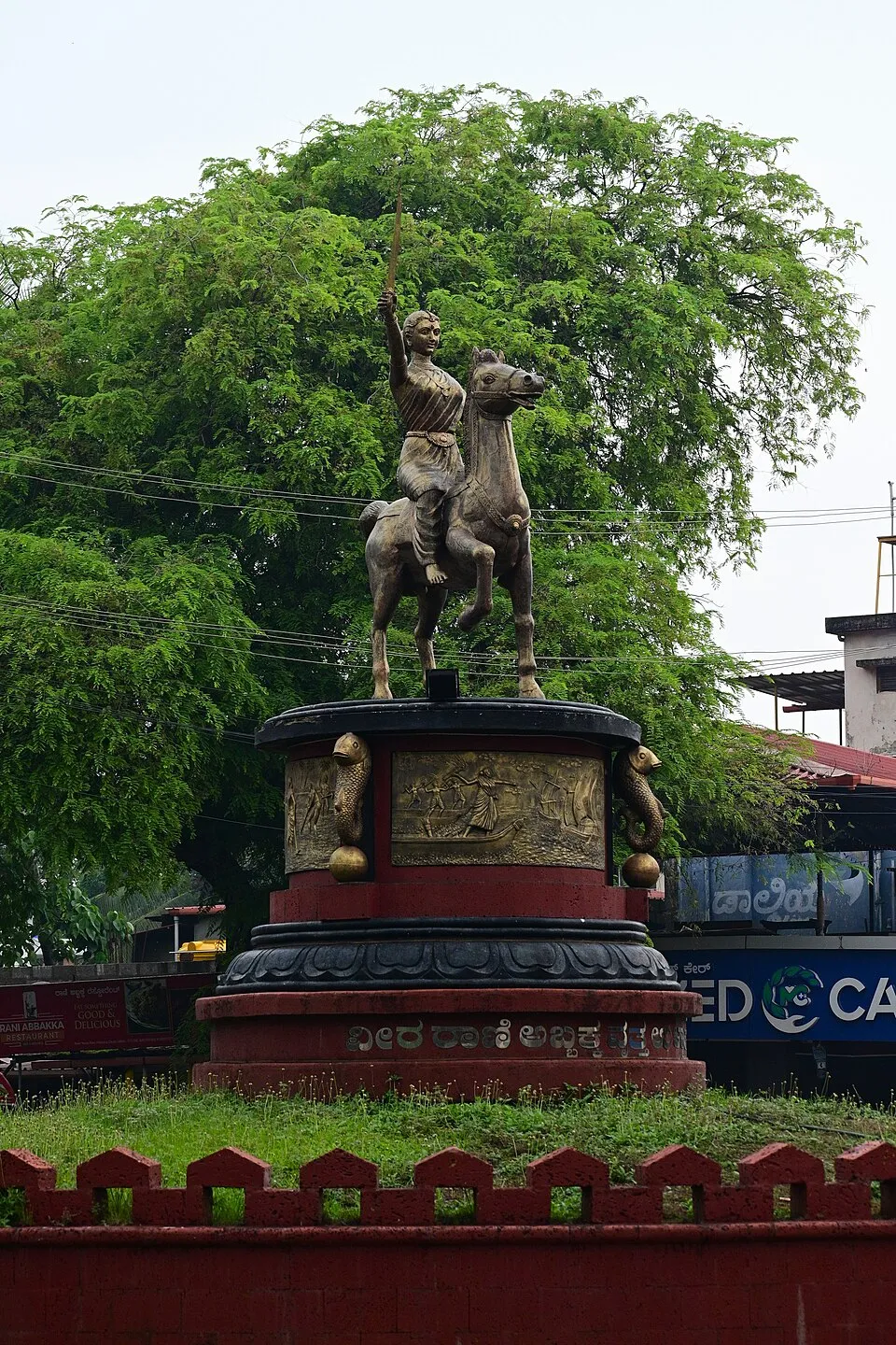 A bronze statue of a woman on a rearing horse, one arm raised, on a round red-and-black pedestal with a carved panel of boats and a Kannada inscription