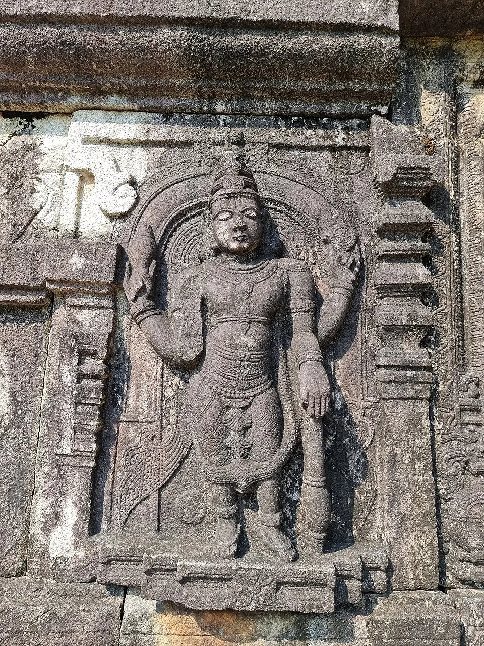 A weathered stone relief of a standing figure with four arms under an arch, carved on the wall of a temple