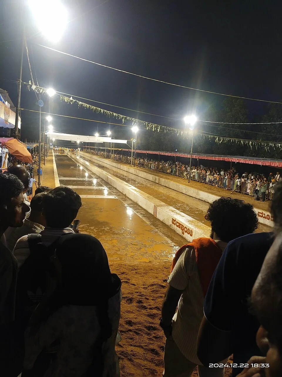 A floodlit kambala track at night at Mulki, a long channel of muddy water between stands of spectators, with bunting overhead