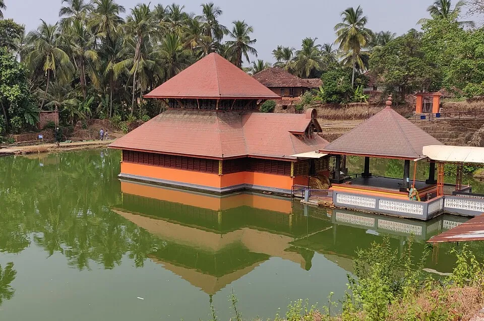 The red-roofed sanctum of the Ananthapura temple and the covered footbridge beside it, standing in the green water of the lake with coconut palms behind
