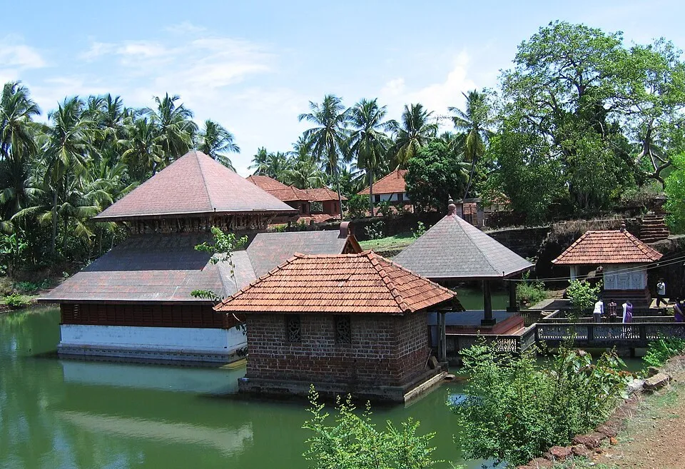 The layered roof of the Ananthapura sanctum seen from another side of the lake, with a small brick and tile shrine on the water in front and tall palms behind