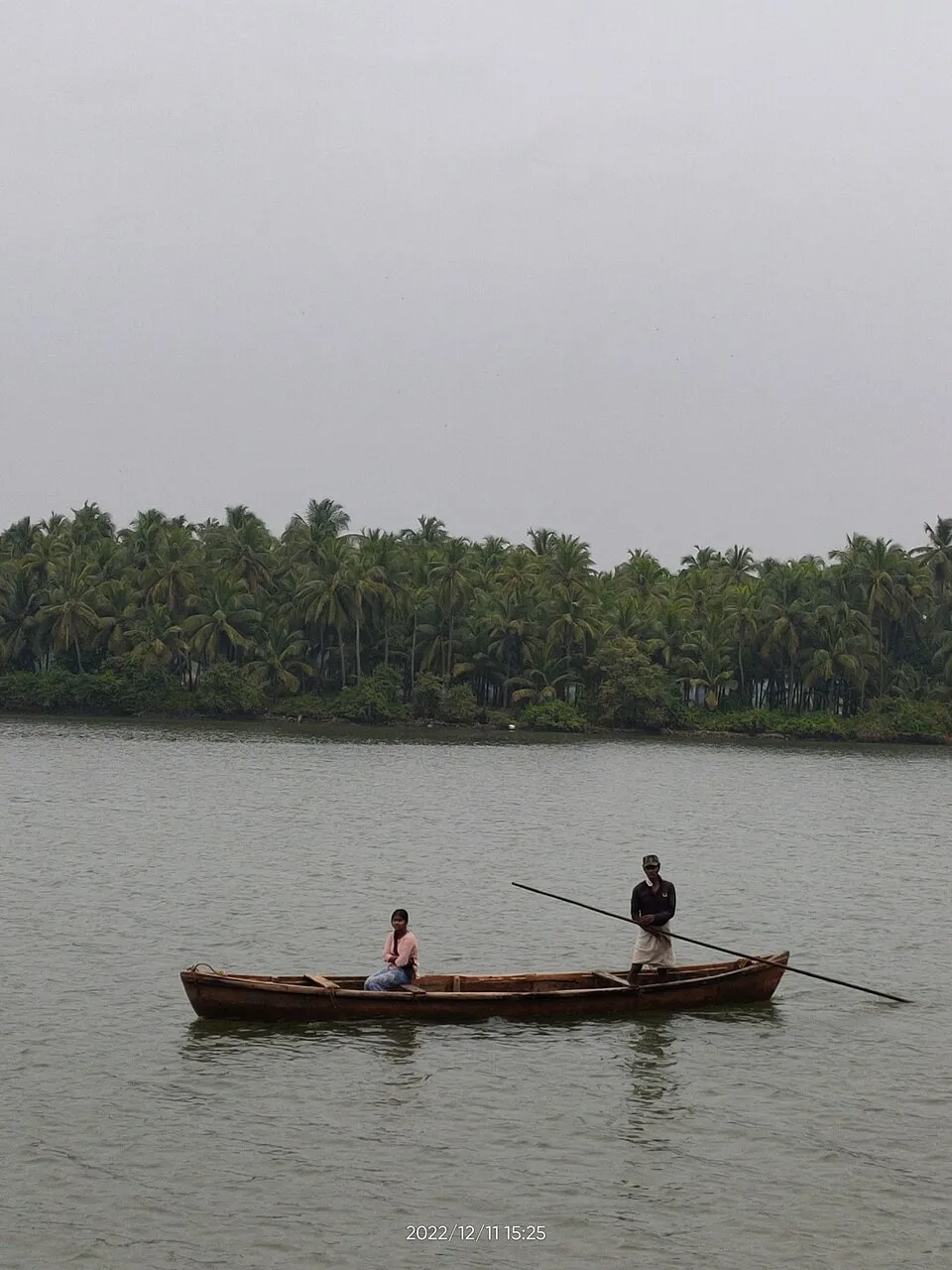 Two men in a wooden boat on the calm Shambhavi backwater near Mulki, with palm-lined banks on the far side