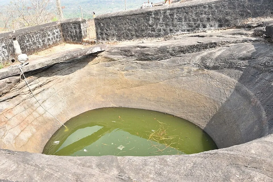A round pond cut into the rock of Karinja hill, holding green water, with a low stone wall above it